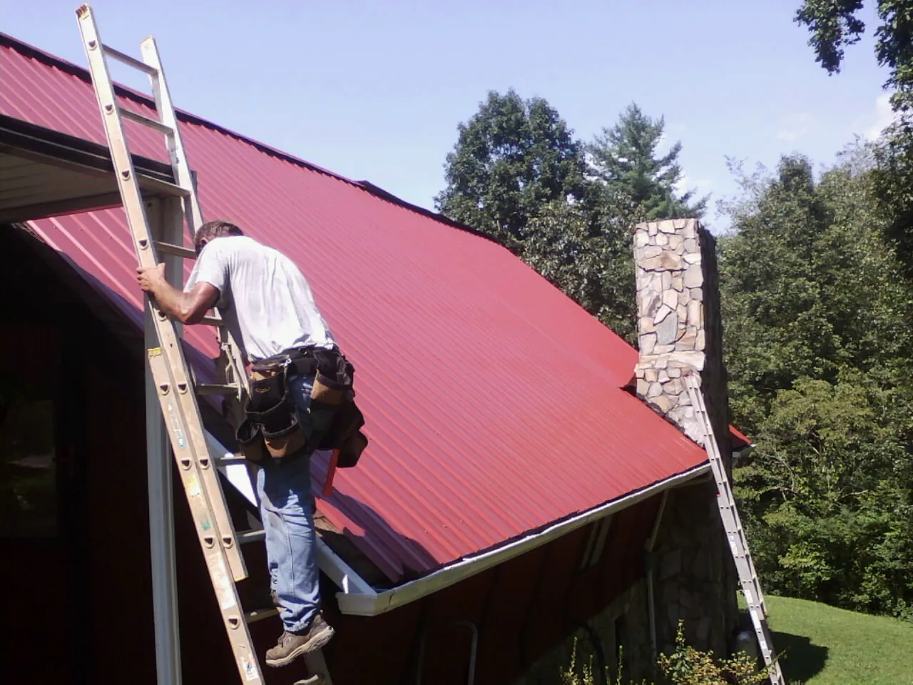 Roofer on a ladder beside a red metal roof and stone chimney — Soffit & Fascia Repair by Roofing Brothers in Waynesville